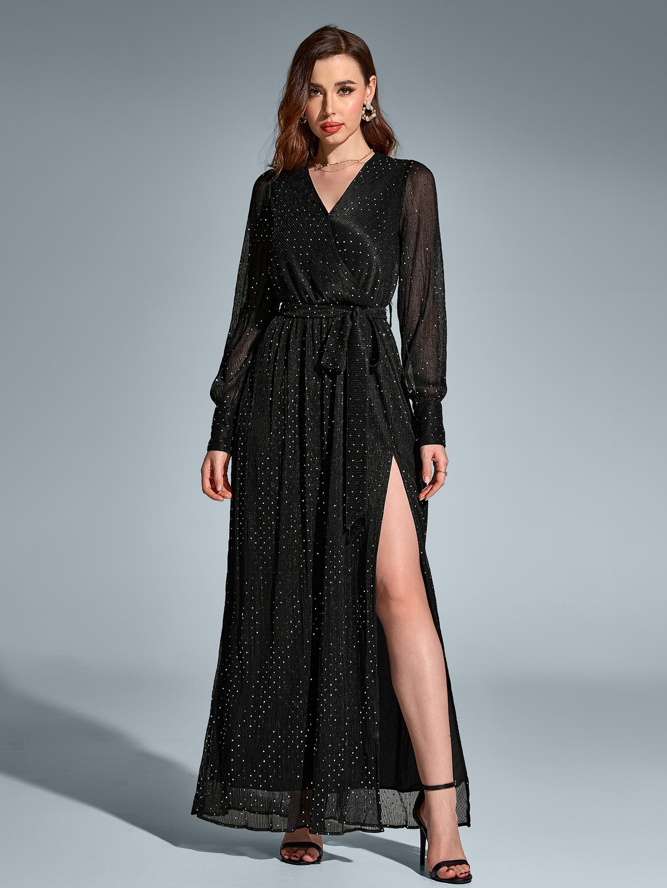 Front view of a woman in a black long-sleeved maxi dress with a v-neck and high slit, adorned with scattered metallic dots, standing with legs apart.