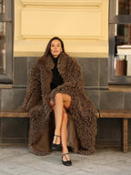 Woman sitting on a bench wearing a long brown faux fur coat.