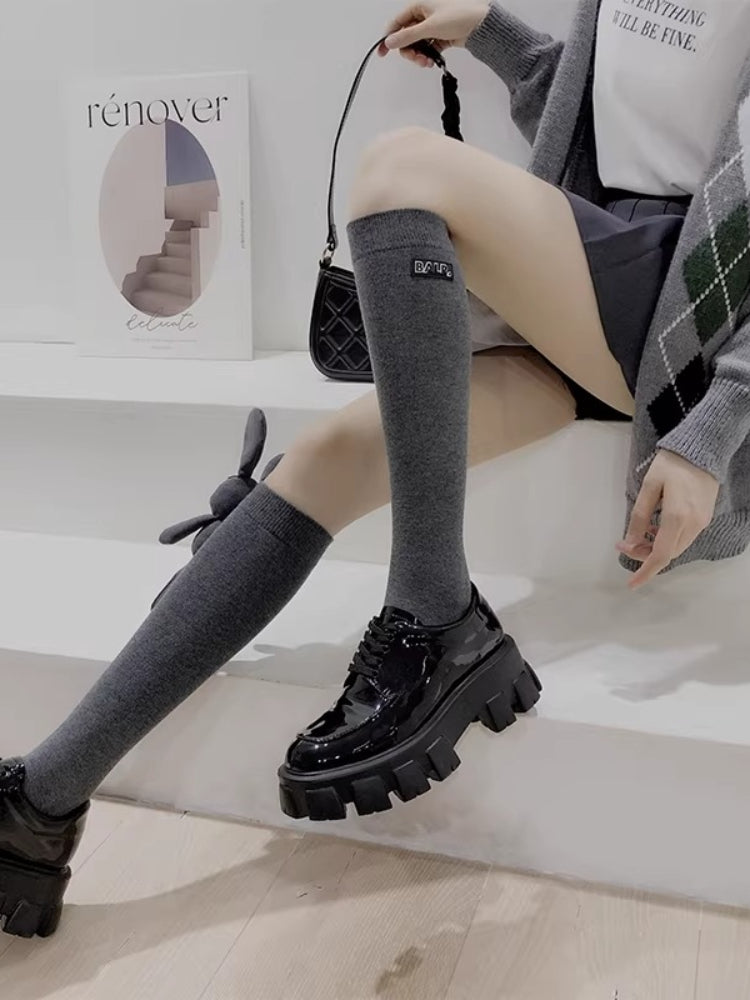 A person sitting, showcasing grey knee-high socks with a bunny charm and black patent leather platform shoes.