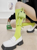 A person wearing lime green knee-high socks with a detachable plush frog toy and white loafers, holding a small green handbag.
