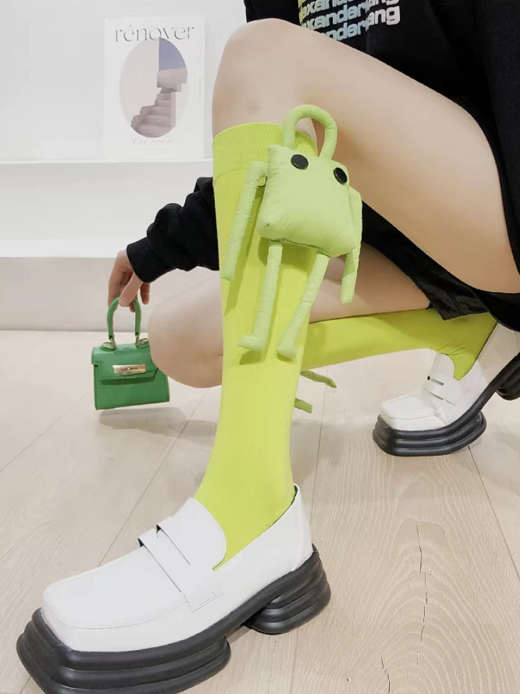 A person wearing lime green knee-high socks with a detachable plush frog toy and white loafers, holding a small green handbag.