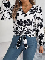 Woman wearing a white long-sleeve blouse with a black floral print and a tie-front detail, adjusting her sleeve.