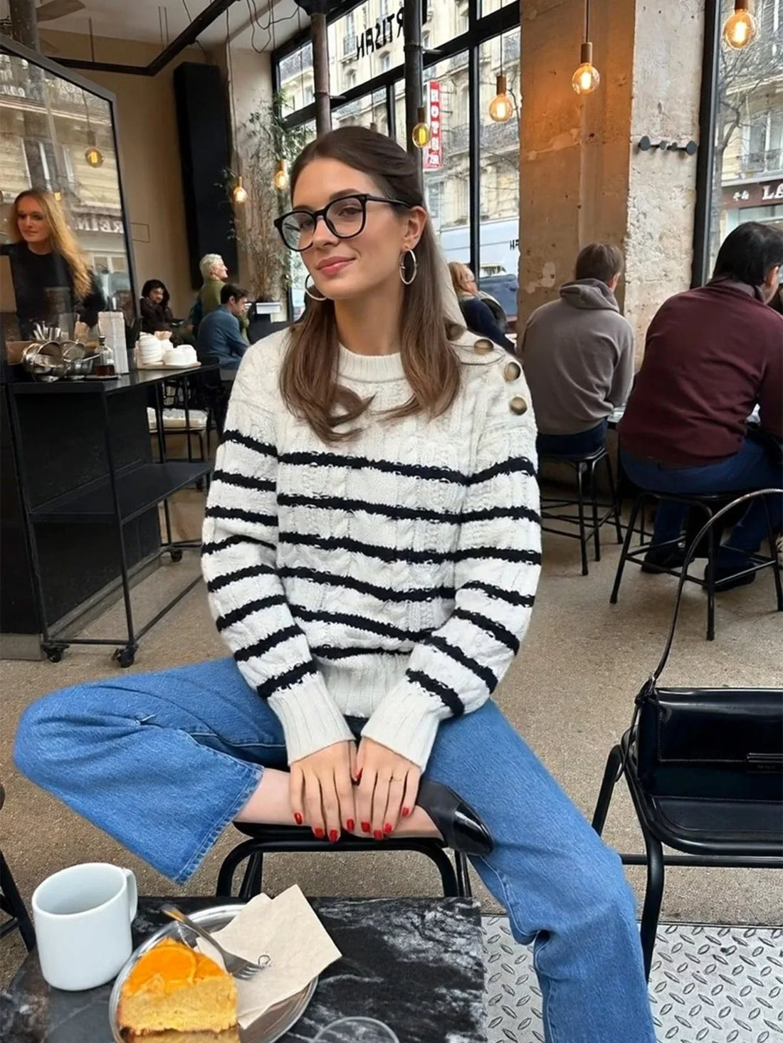 Woman wearing a white and black striped knit sweater with button details on the shoulder, blue denim jeans, and black ballet flats, sitting in a cafe.
