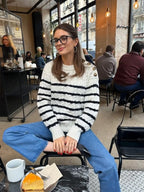 Woman wearing a white and black striped knit sweater with button details on the shoulder, blue denim jeans, and black ballet flats, sitting in a cafe.