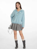 Full-body shot of a model wearing a light blue fuzzy knit collared v-neck sweater with a ribbed hem, paired with a grey pleated mini skirt, grey boots, and a silver handbag.