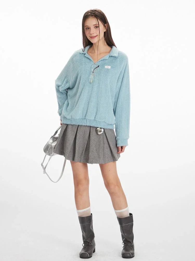 Full-body shot of a model wearing a light blue fuzzy knit collared v-neck sweater with a ribbed hem, paired with a grey pleated mini skirt, grey boots, and a silver handbag.