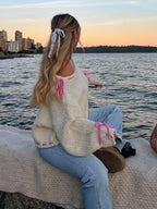 Woman sitting by the water at sunset, wearing a cream knit sweater with pink tie details on the sleeves.
