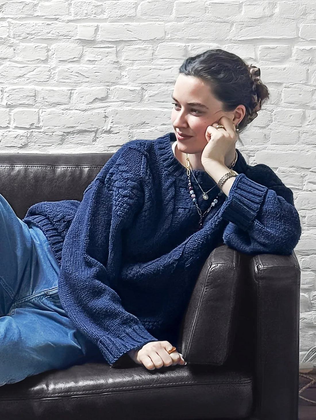 Woman sitting on a brown leather couch wearing a navy blue cable knit sweater and light wash jeans.