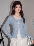 Woman wearing a light blue knit cardigan with white bow tie closures over a white top and a white tiered skirt.