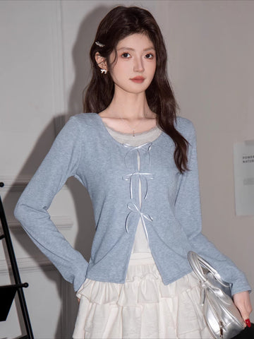 Woman wearing a light blue knit cardigan with white bow tie closures over a white top and a white tiered skirt.
