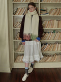 Woman wearing a cream-colored scarf with fringed ends transitioning to pink and green, a dark maroon crewneck sweater, a white tiered lace-trimmed skirt, and white sneakers.