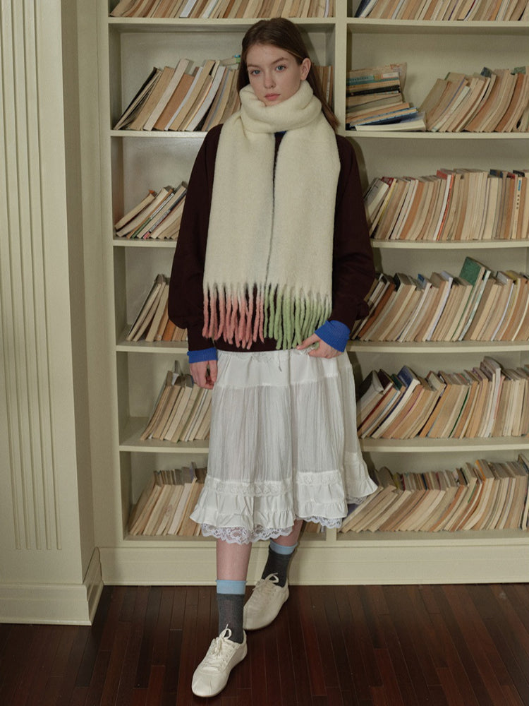 Woman wearing a cream-colored scarf with fringed ends transitioning to pink and green, a dark maroon crewneck sweater, a white tiered lace-trimmed skirt, and white sneakers.
