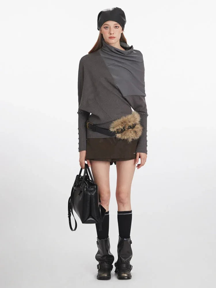Woman wearing a grey asymmetrical knit sweater with draped overlay, dark shorts, black boots, black beanie, and carrying a black handbag.