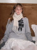Woman sitting on a bed wearing a grey sweatshirt and cream scarf