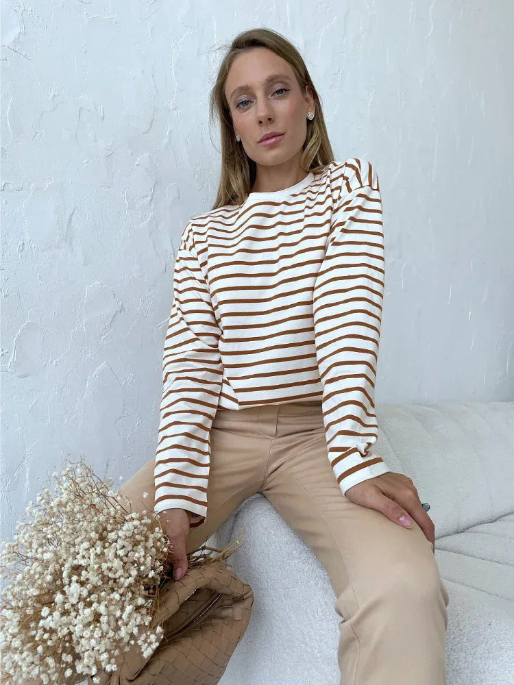 Woman sitting on a couch wearing a white and brown striped long sleeve top and beige pants, holding a bouquet of dried flowers and a woven handbag.