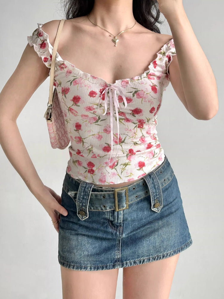 Woman wearing a white floral print top with ruffled off-the-shoulder cap sleeves and a sweetheart neckline with a pink ribbon tie, accessorized with a pink handbag and denim skirt.