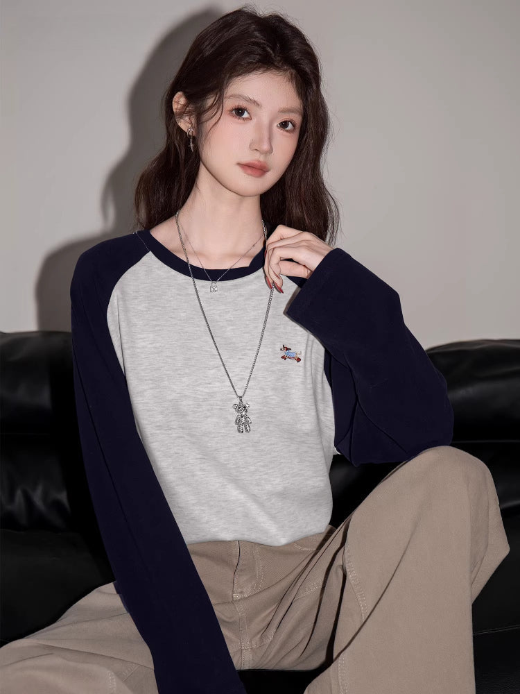 Close-up of a woman wearing a light grey long-sleeve raglan shirt with navy blue sleeves and a small embroidered bird graphic.