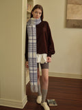 Woman posing with one hand on a wooden table, wearing a white vest over a white long-sleeved shirt, a white pleated skirt, grey polka dot tights, silver boots, and a grey plaid scarf.