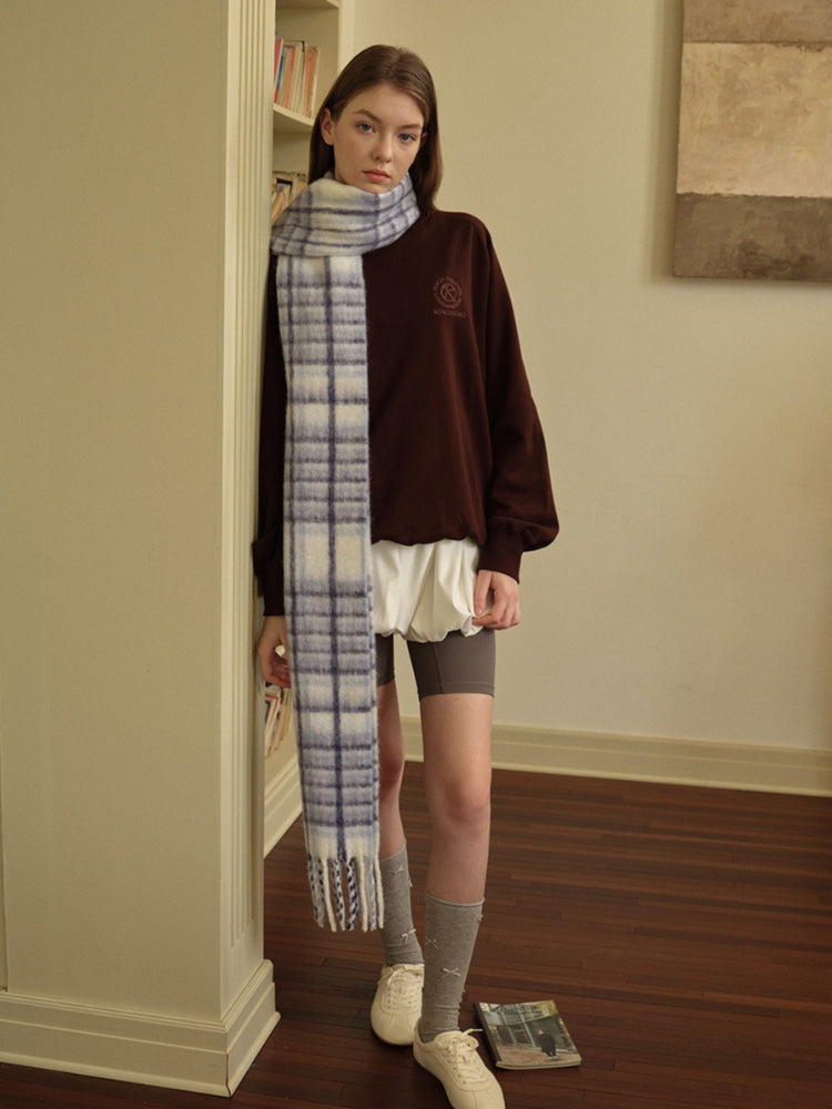 Woman posing with one hand on a wooden table, wearing a white vest over a white long-sleeved shirt, a white pleated skirt, grey polka dot tights, silver boots, and a grey plaid scarf.