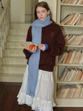 Woman holding a book and wearing a brown velour sweatshirt with a light blue scarf, white ruffled skirt, grey socks, and white sneakers, standing in front of a bookshelf.