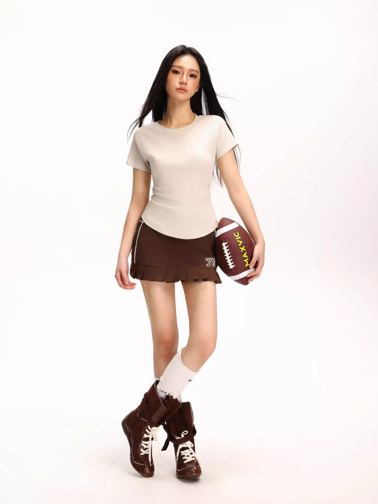 Woman in a beige fitted short-sleeve t-shirt, brown pleated skirt, and knee-high socks holding a football