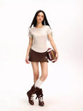 Woman in a beige fitted short-sleeve t-shirt, brown pleated skirt, and knee-high socks holding a football