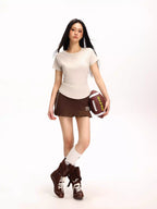 Woman in a beige fitted short-sleeve t-shirt, brown pleated skirt, and knee-high socks holding a football