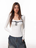 Woman posing with hands in pockets, wearing a white ribbed long-sleeve top with a black bow and distressed denim shorts.