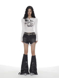 Full body shot of a woman in a white long-sleeve graphic print cropped top, distressed denim shorts, and oversized leg warmers.