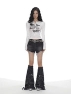 Full body shot of a woman in a white long-sleeve graphic print cropped top, distressed denim shorts, and oversized leg warmers.