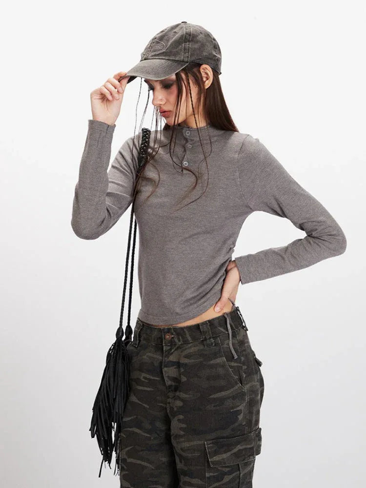 Close-up of a model's upper body wearing a grey long-sleeve mock neck cropped top with button details, and camouflage cargo pants. Model is wearing a grey baseball cap.