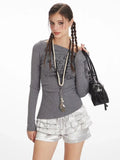 Close-up of a woman wearing a grey long-sleeve off-the-shoulder top with graphic print and layered ruffled silver shorts, accessorized with a long necklace.