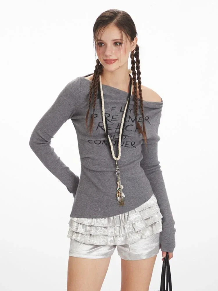 Front view of a woman wearing a grey long-sleeve off-the-shoulder top with graphic print and layered ruffled silver shorts, with braids.