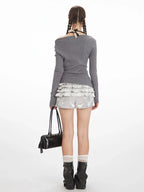 Back view of a woman wearing a grey long-sleeve off-the-shoulder top with graphic print and layered ruffled silver shorts.