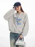 Close-up of a woman wearing a grey graphic print sweatshirt with a cut-out neckline and blue text, posing with her hand on her head.
