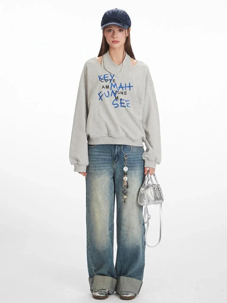 Woman wearing a grey graphic print sweatshirt with a cut-out neckline and blue text, paired with distressed wide-leg jeans and a silver handbag.