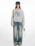 Woman wearing a grey graphic print sweatshirt with a cut-out neckline and blue text, paired with distressed wide-leg jeans and a silver handbag.