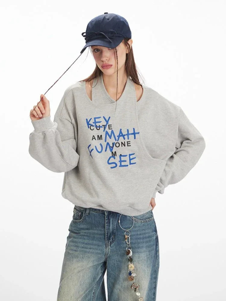 Woman wearing a grey graphic print sweatshirt with a cut-out neckline and blue text, holding a string from a navy blue cap.