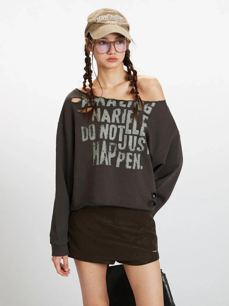 Close-up front view of a woman wearing a dark grey distressed graphic print cropped sweatshirt with an off-the-shoulder neckline, showing graphic print detail.