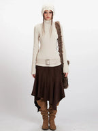 Woman wearing a cream ribbed knit turtleneck sweater, brown asymmetrical hem skirt, cream knit beanie, and brown suede boots.