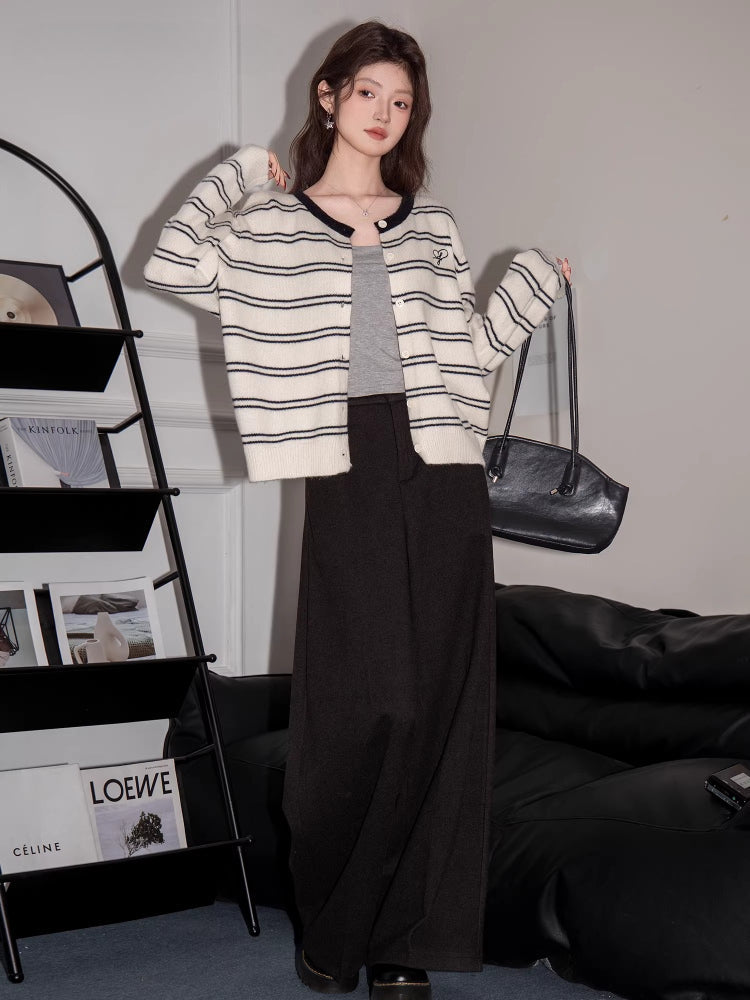 Full body shot of a woman wearing a striped knit cardigan over a grey tank top and dark trousers.