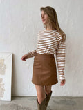 Woman wearing a white and brown striped long sleeve top and a brown faux leather mini skirt with cowboy boots.