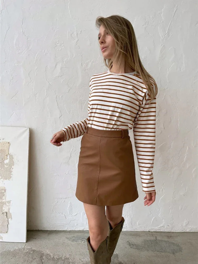 Woman wearing a white and brown striped long sleeve top and a brown faux leather mini skirt with cowboy boots.