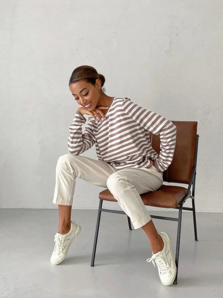 Woman sitting on a chair wearing a striped long-sleeve top and cream trousers, with a thoughtful expression.