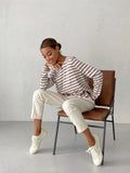 Woman sitting on a chair wearing a striped long-sleeve top and cream trousers, with a thoughtful expression.