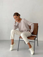 Woman sitting on a chair wearing a striped long-sleeve top and cream trousers, with a thoughtful expression.