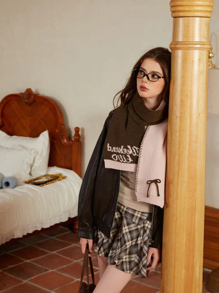 Woman wearing a light pink knit scarf with dark trim and embroidered text, sitting on a bed.
