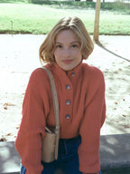 Front view of a woman wearing a coral knit sweater with a high neckline and button placket, outdoors.