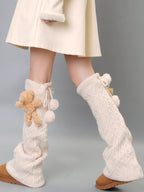 Cream cable knit leg warmers with teddy bear and pom-pom details, worn with a cream dress and tan boots.
