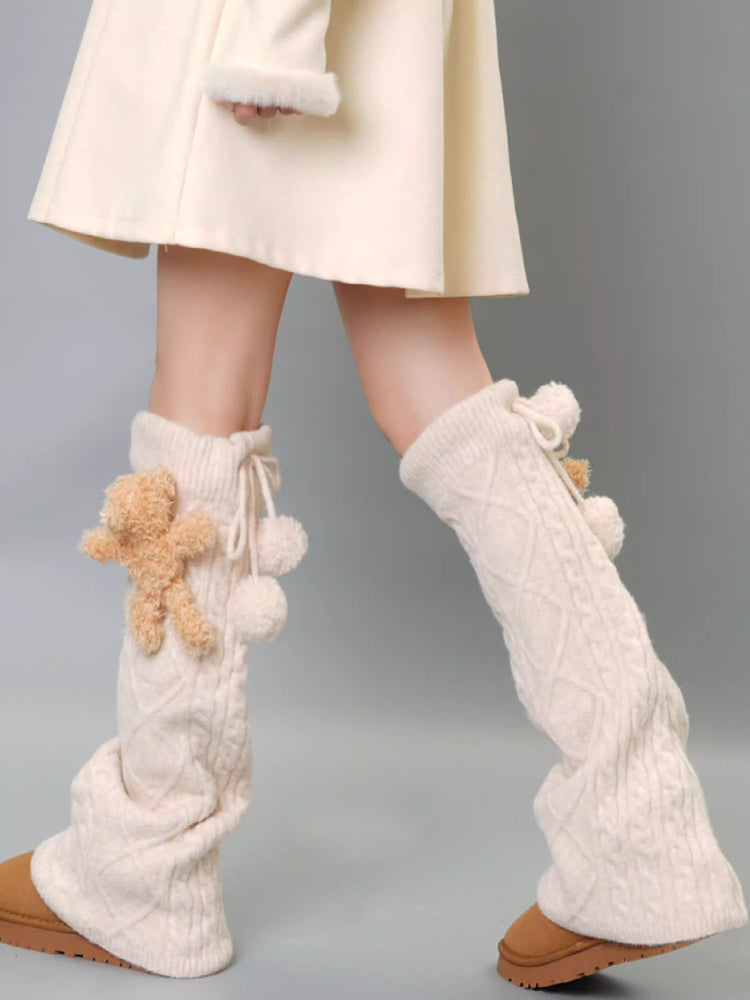 Cream cable knit leg warmers with teddy bear and pom-pom details, worn with a cream dress and tan boots.